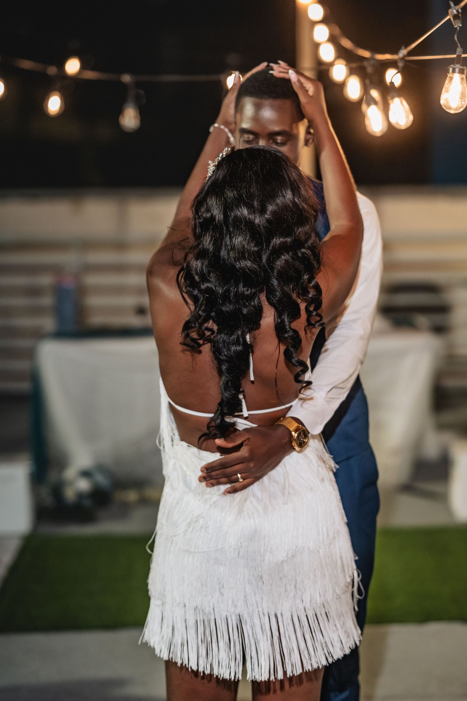 Couple dancing outdoors at night under string lights, woman in white fringe dress with curly hair, man in blue suit.