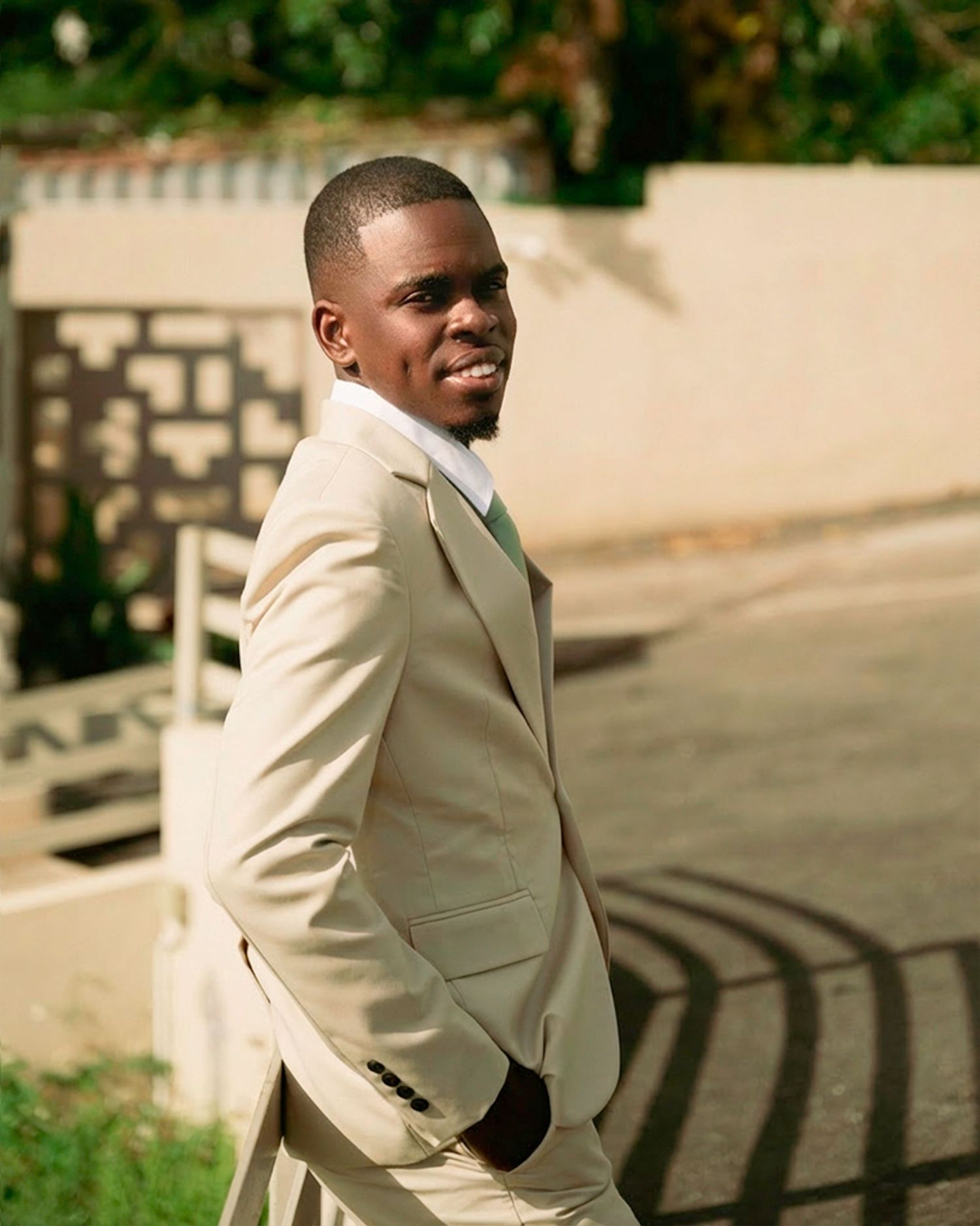 A young man in a cream suit smiles over his shoulder outdoors.