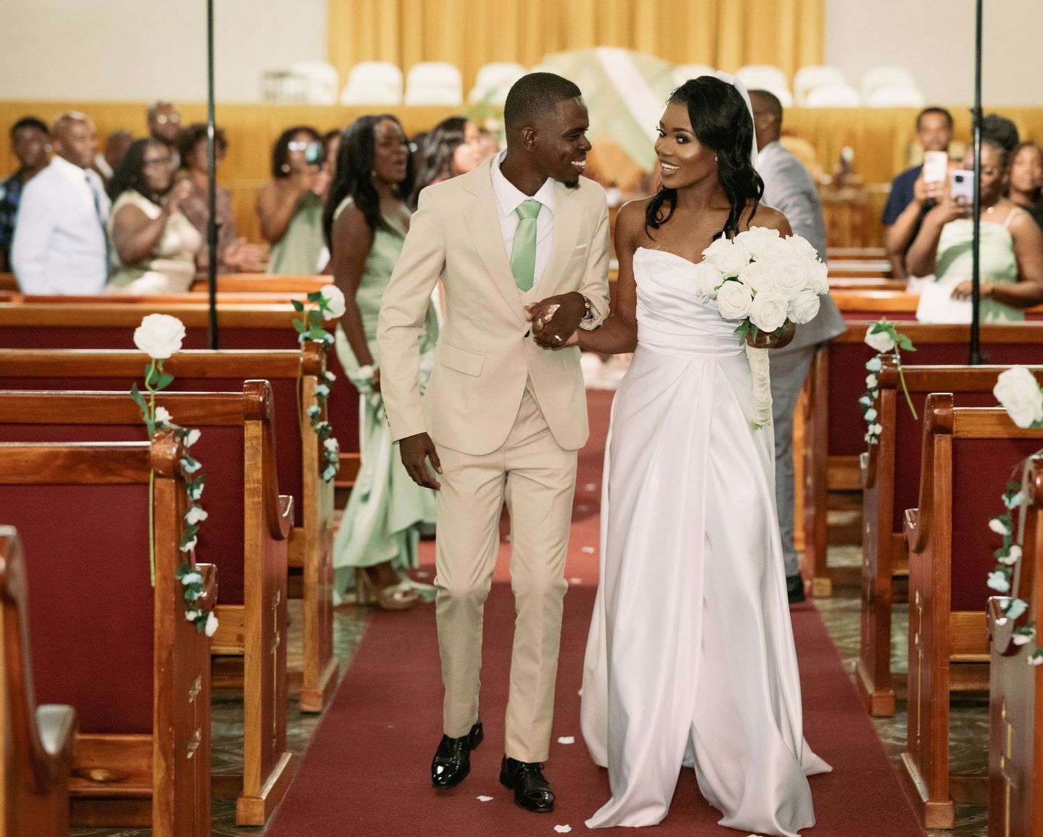 Newlyweds walk hand in hand down church aisle after ceremony, groom in beige suit, bride in white gown.