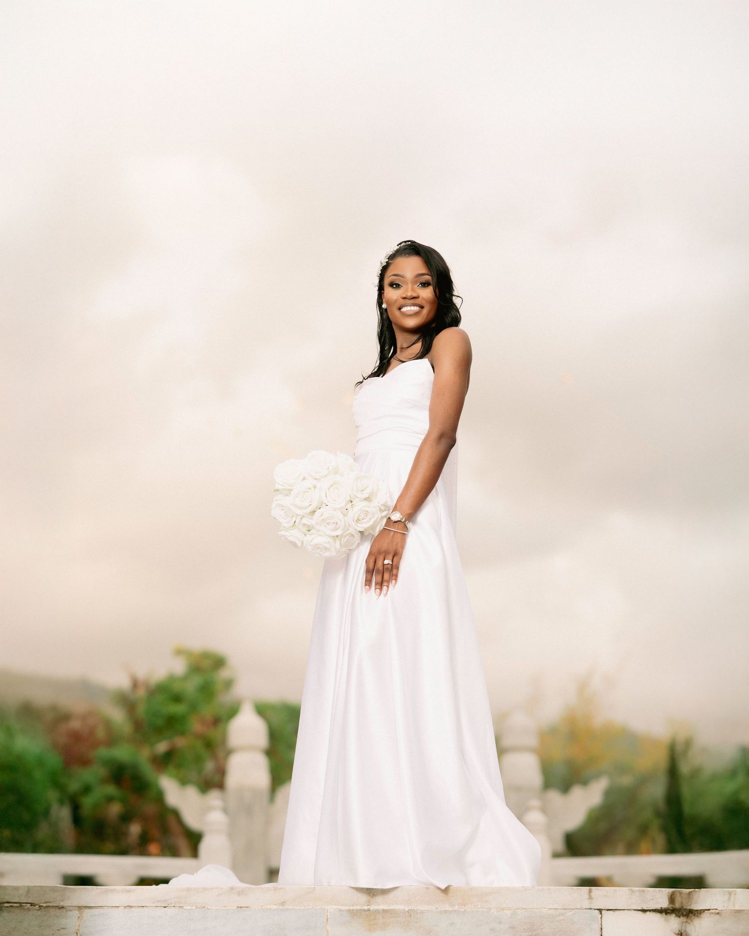 Smiling Black bride in elegant white gown holding white rose bouquet, standing on steps against cloudy sky backdrop.