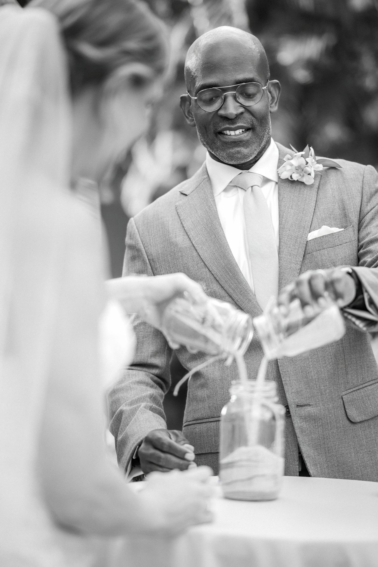 Black and white photo of a groom smiling during a sand unity ceremony at an outdoor wedding.