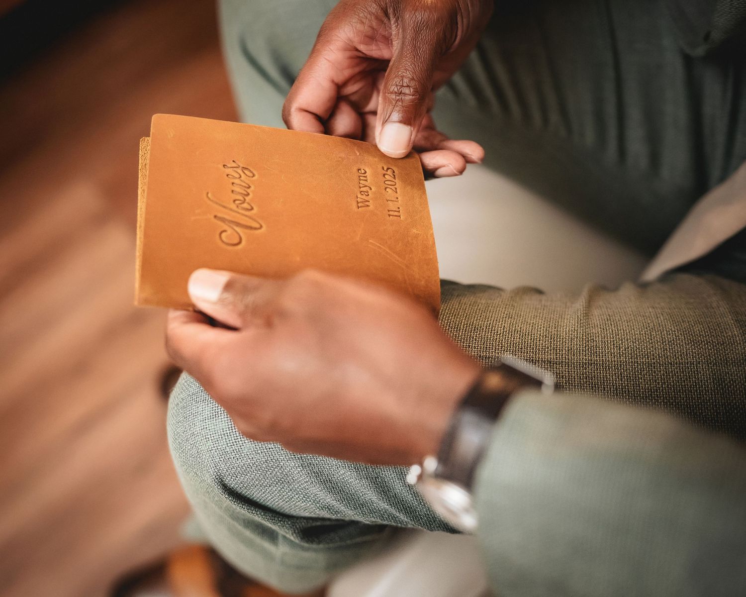 Man holding a tan leather journal with embossed script lettering, wearing a watch and sage green suit.