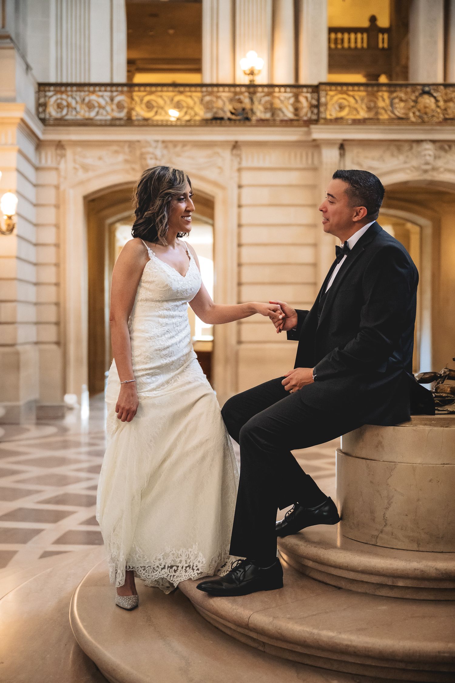 Bride and groom holding hands at San Francisco City Hall, she in lace gown, he in black tuxedo.