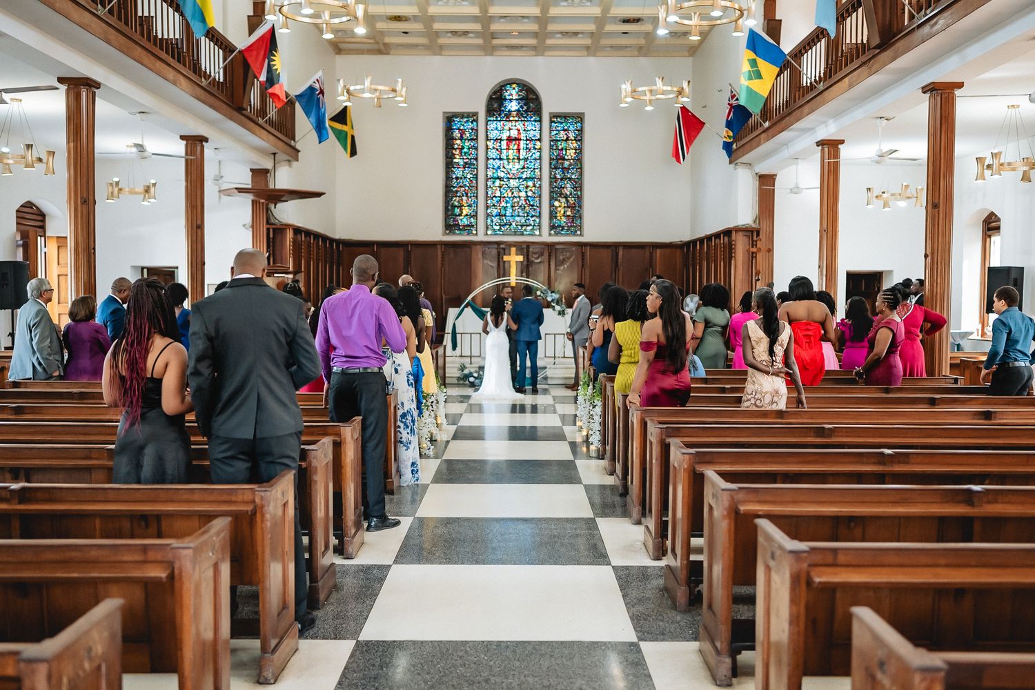 Guests standing in a beautiful church with stained glass windows and colorful flags during a wedding ceremony.