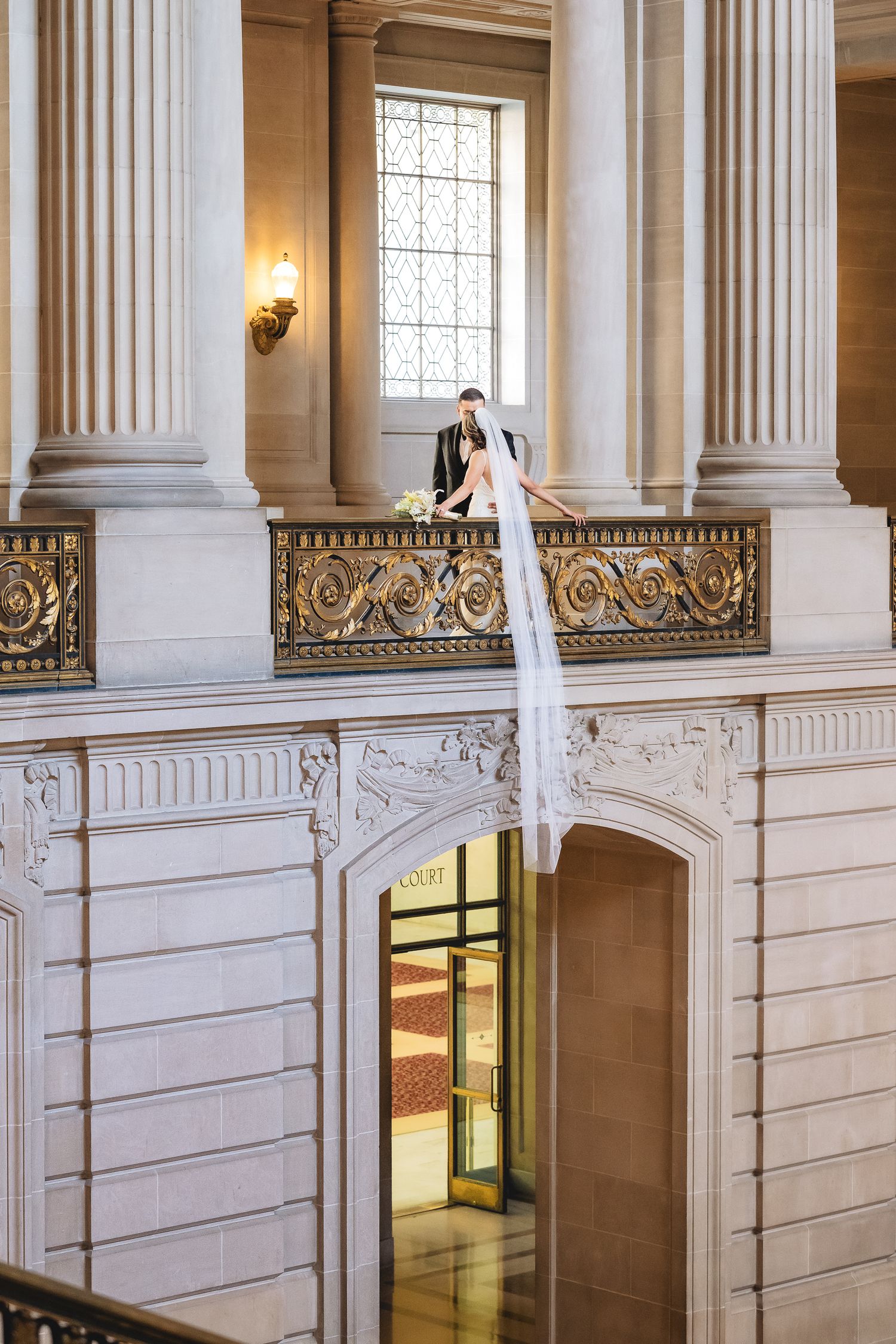 Bride and groom share a romantic moment on a grand balcony at San Francisco City Hall, with a flowing white veil cascading over the railing.