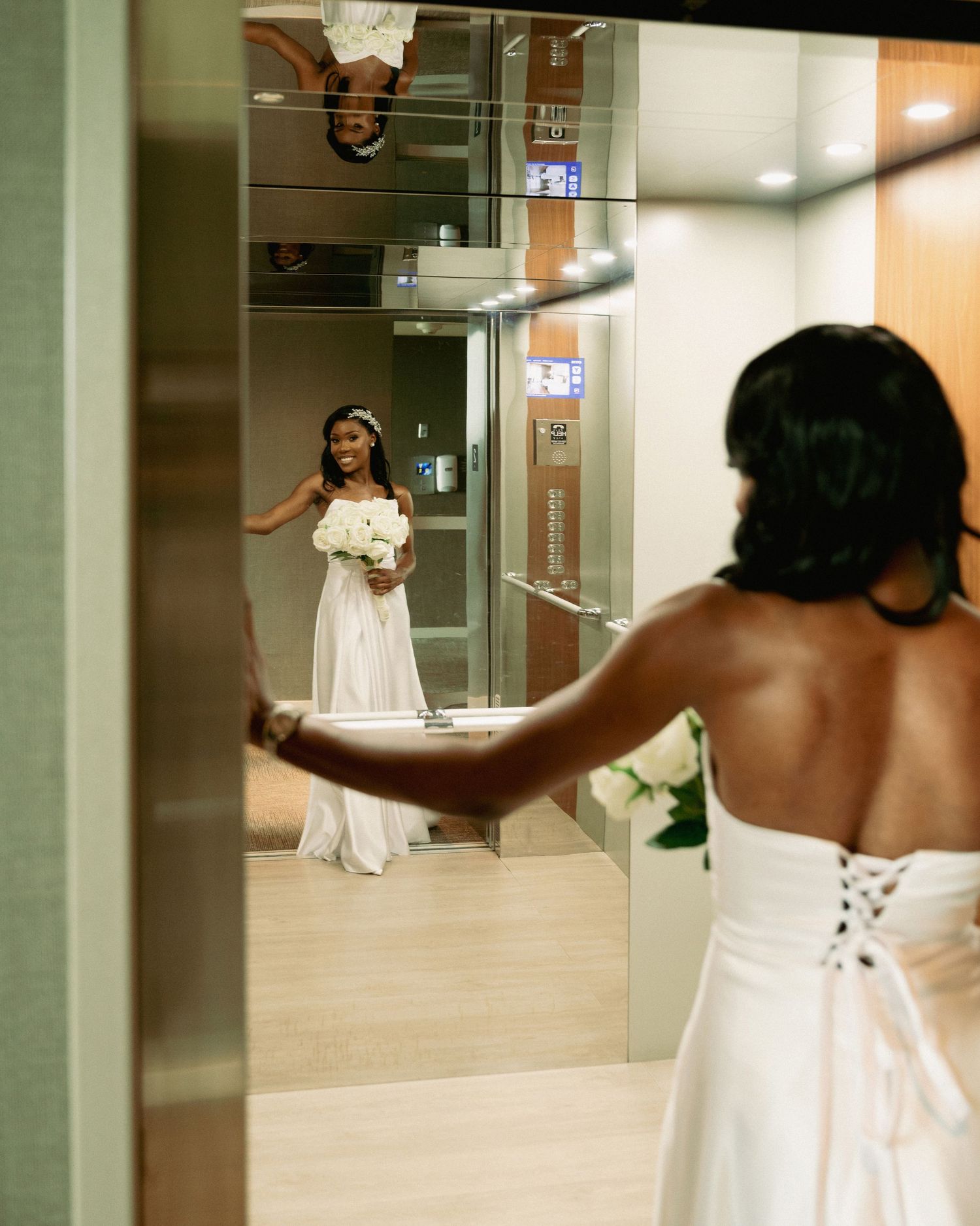 Bride in white gown holding bouquet reflected in elevator mirror at her wedding venue.