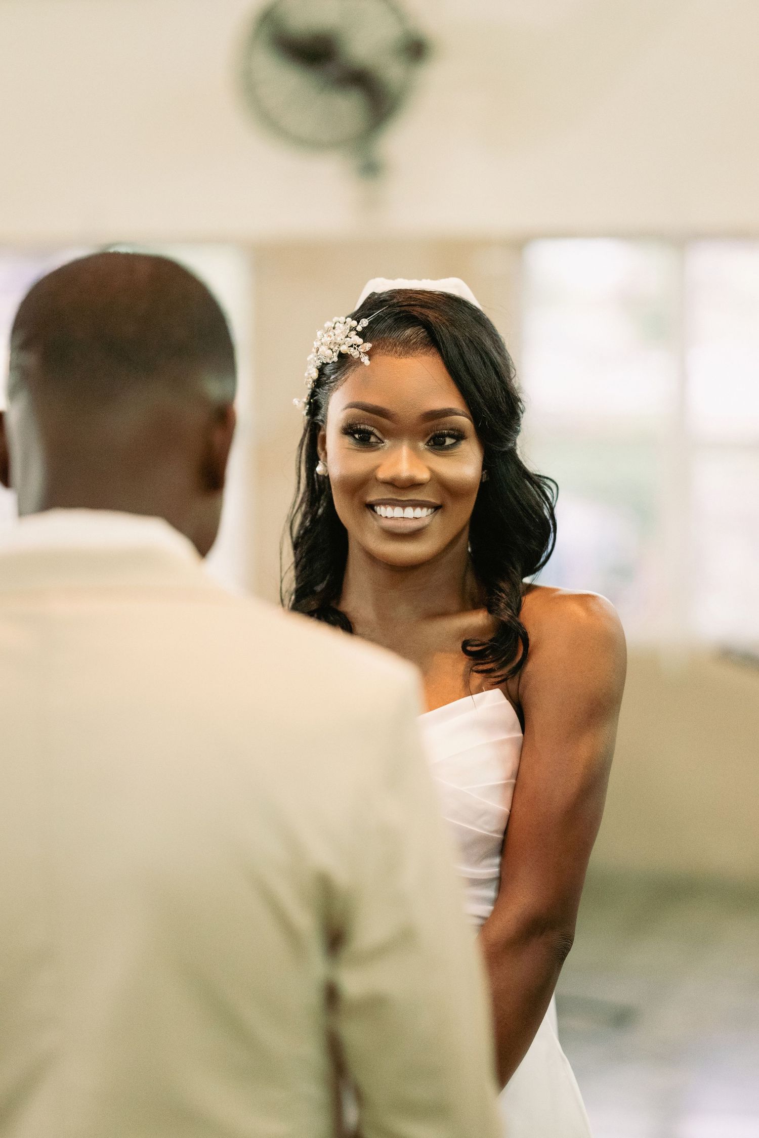 Radiant bride in white strapless dress smiles at groom during wedding ceremony indoors.