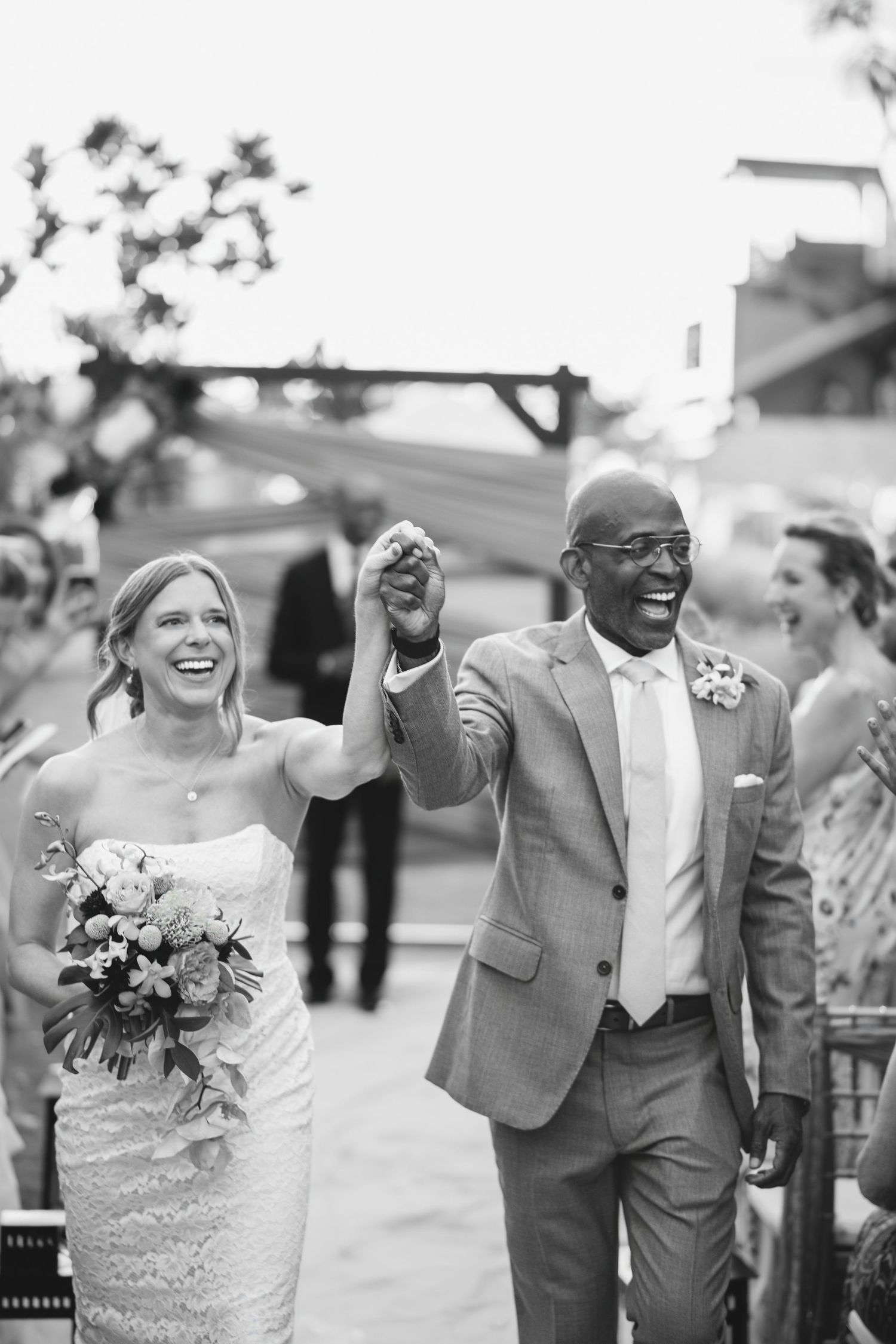 Black and white photo of joyful interracial newlyweds walking down the aisle, bride holding bouquet, groom in gray suit.
