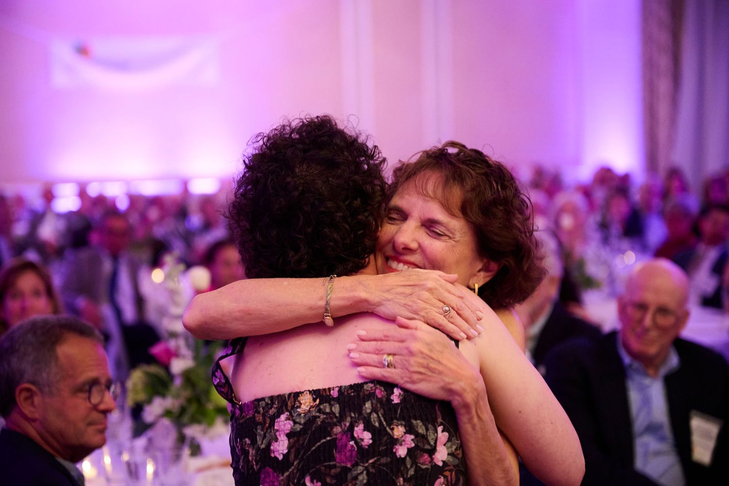 A group of people embrace and dance at an elegant event with purple lighting and formal attire.