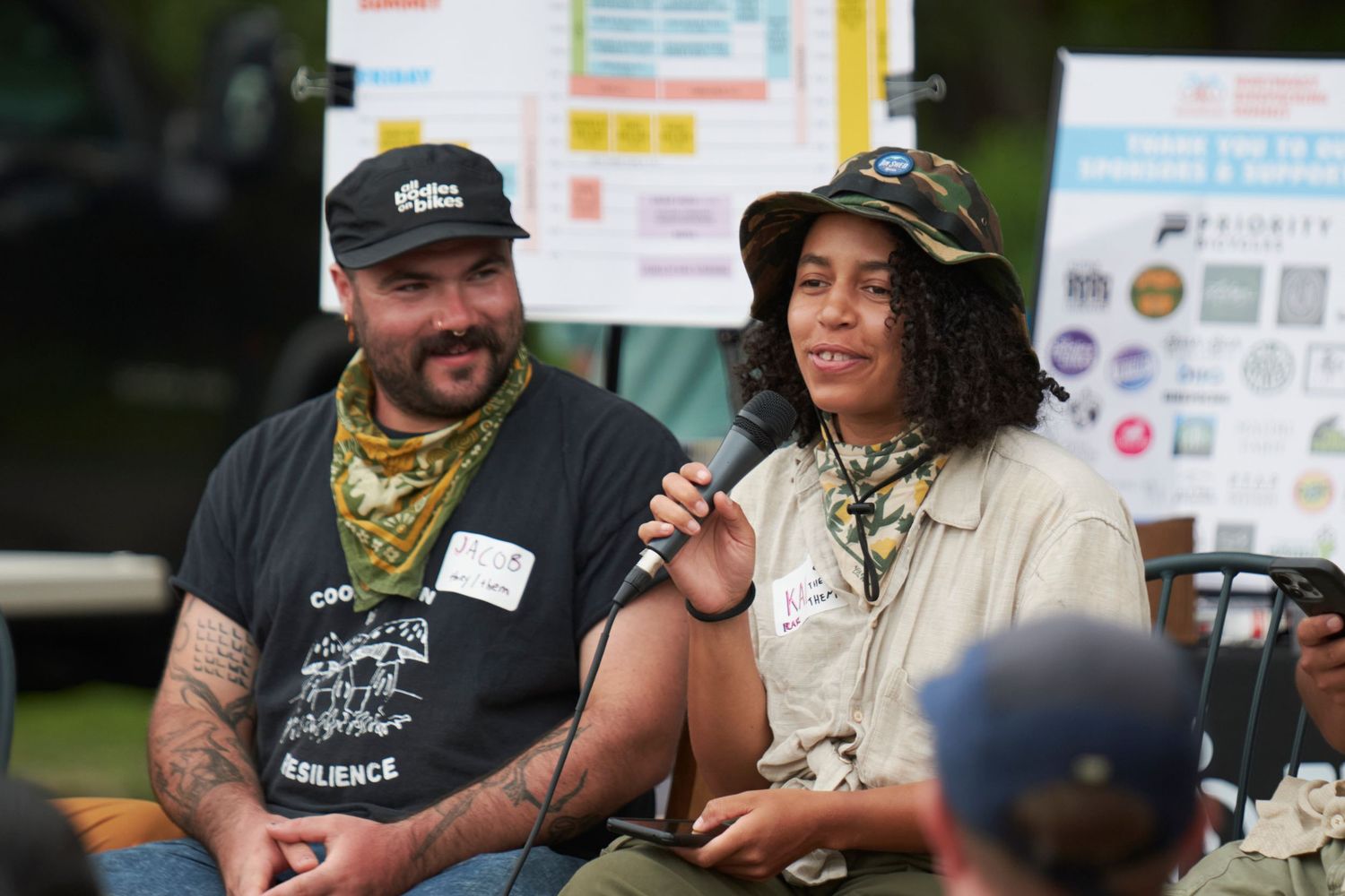 People sit together at an outdoor gathering wearing casual attire and name tags while engaged in conversation.