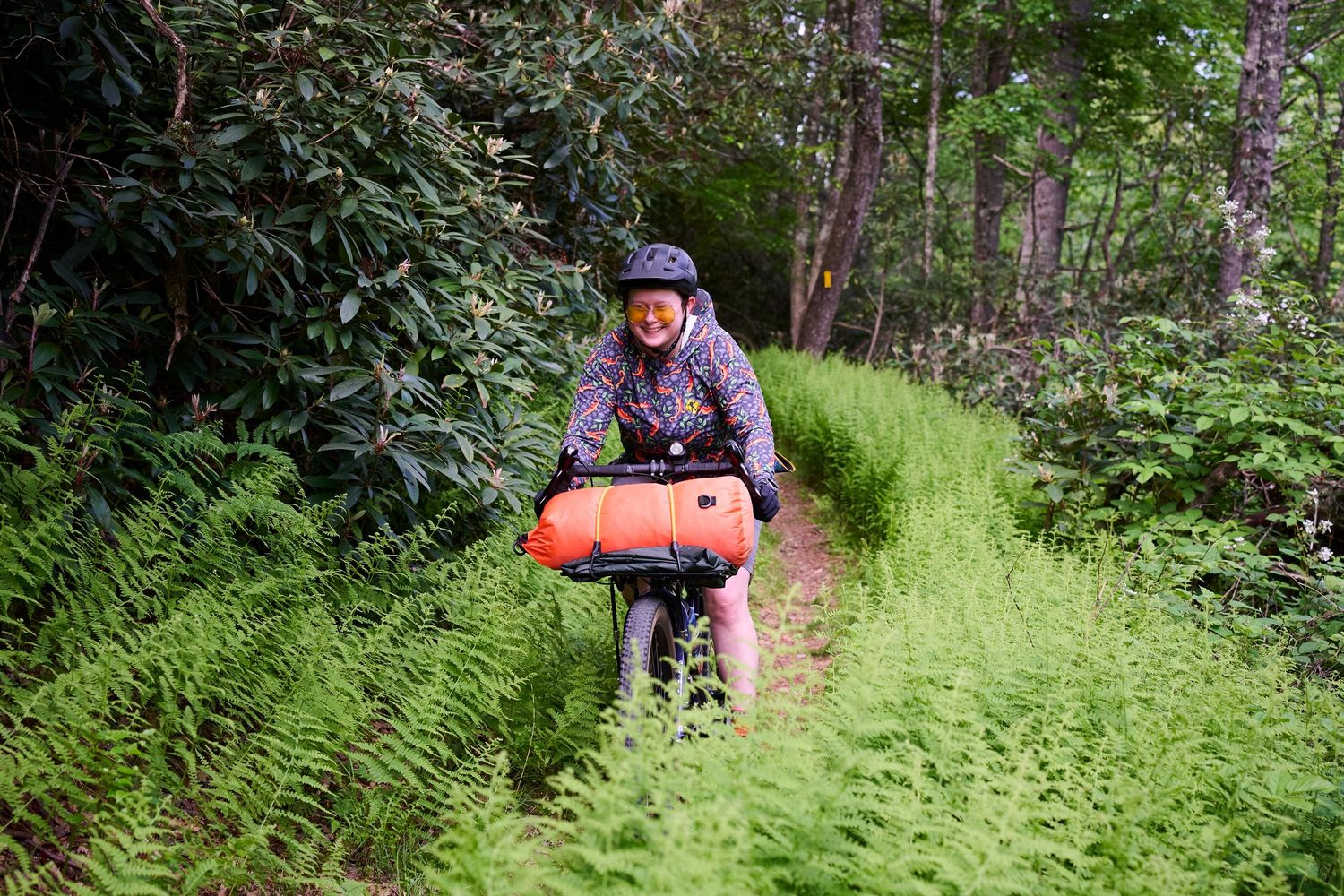 Cyclist riding a bike with orange bag through a lush green fern-lined forest trail wearing a helmet.