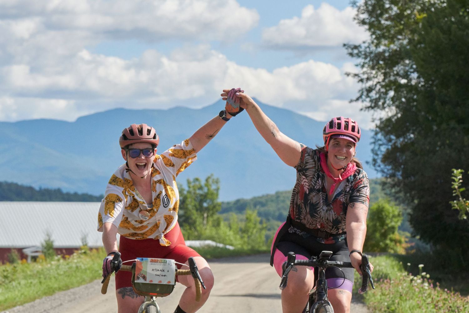 Two cyclists high-five while riding on a scenic mountain road with blue skies and mountains in background.