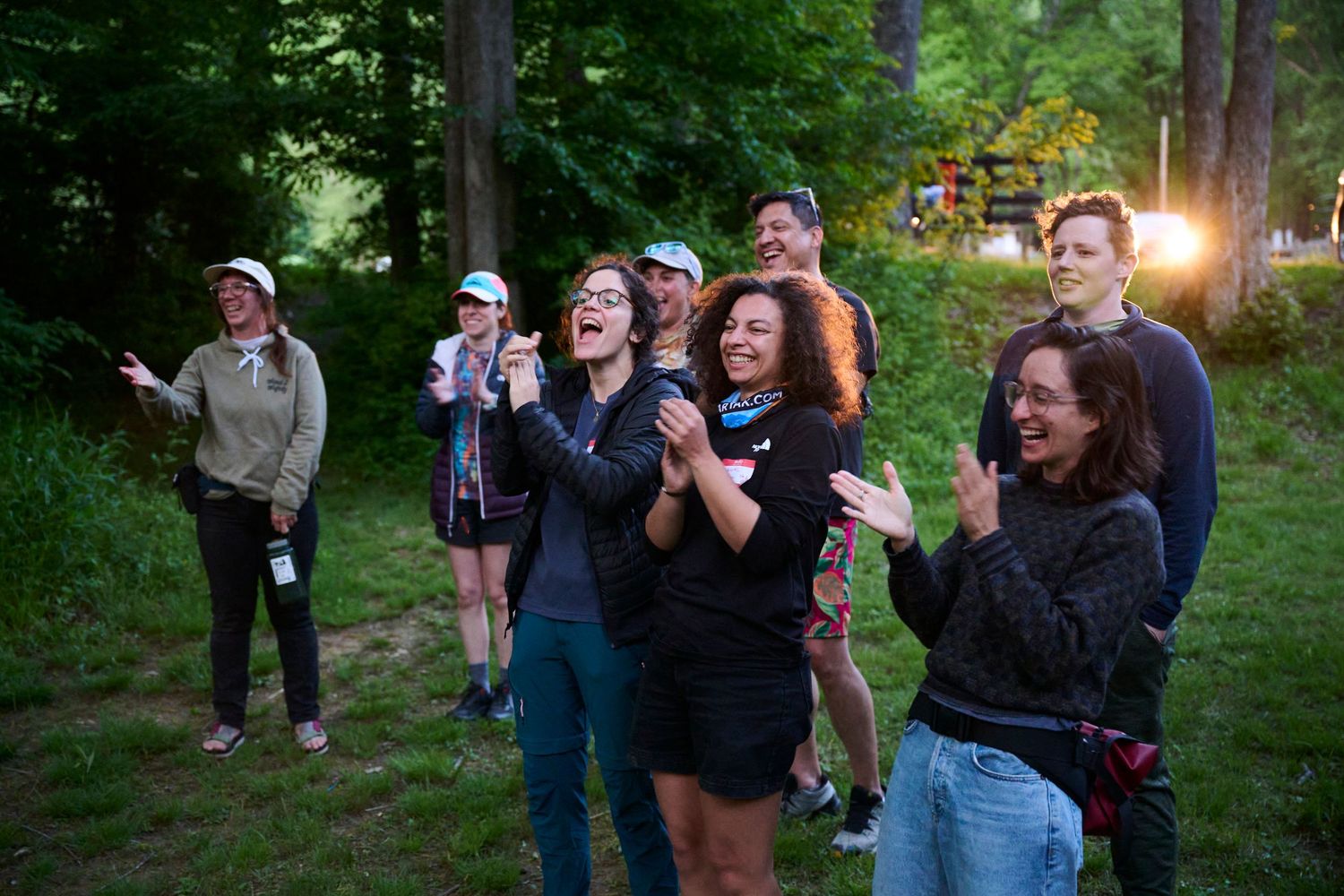Group of people laughing and clapping outdoors in a forest setting during evening gathering.