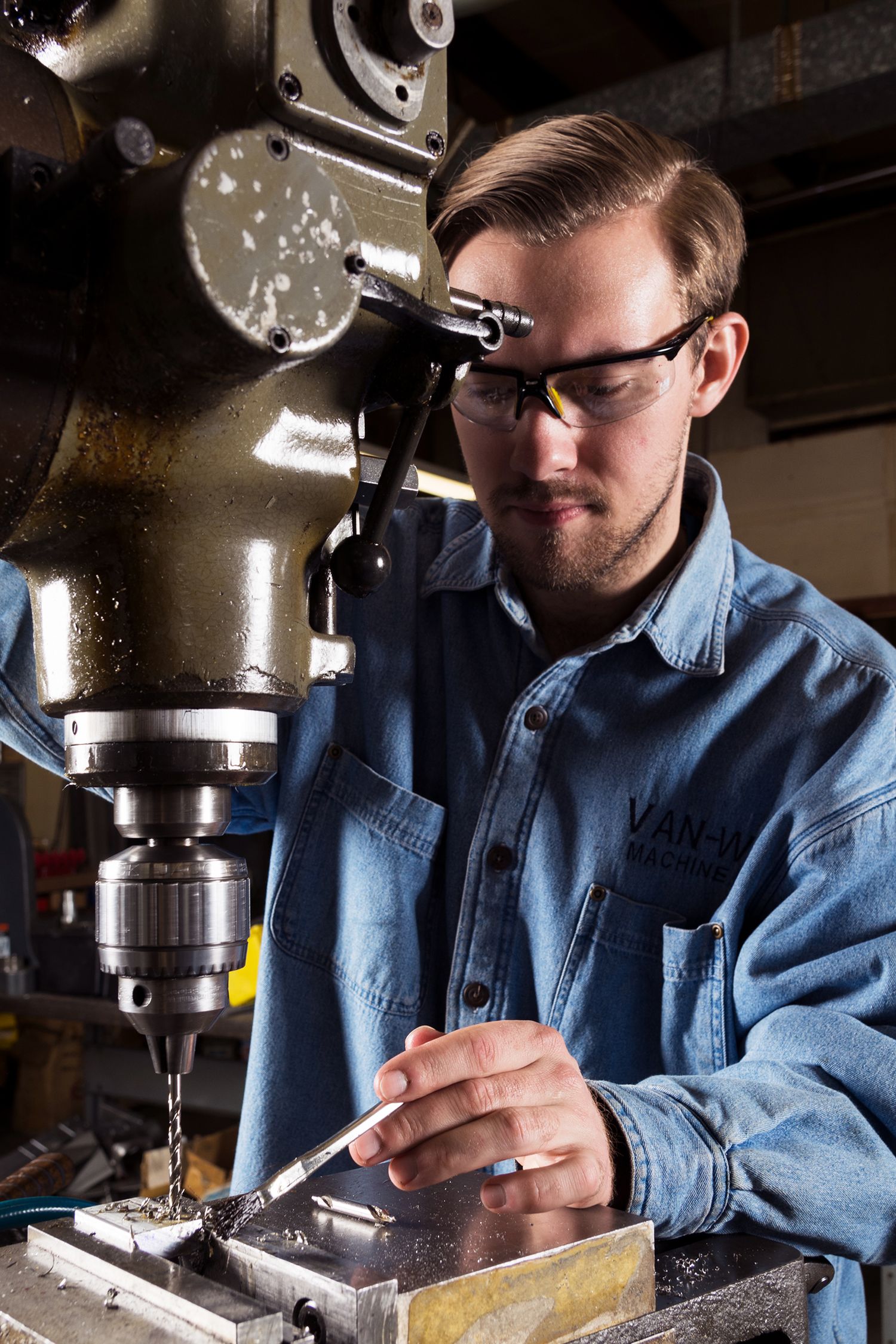 Worker in blue denim jacket uses precision drilling machine in industrial workshop setting.