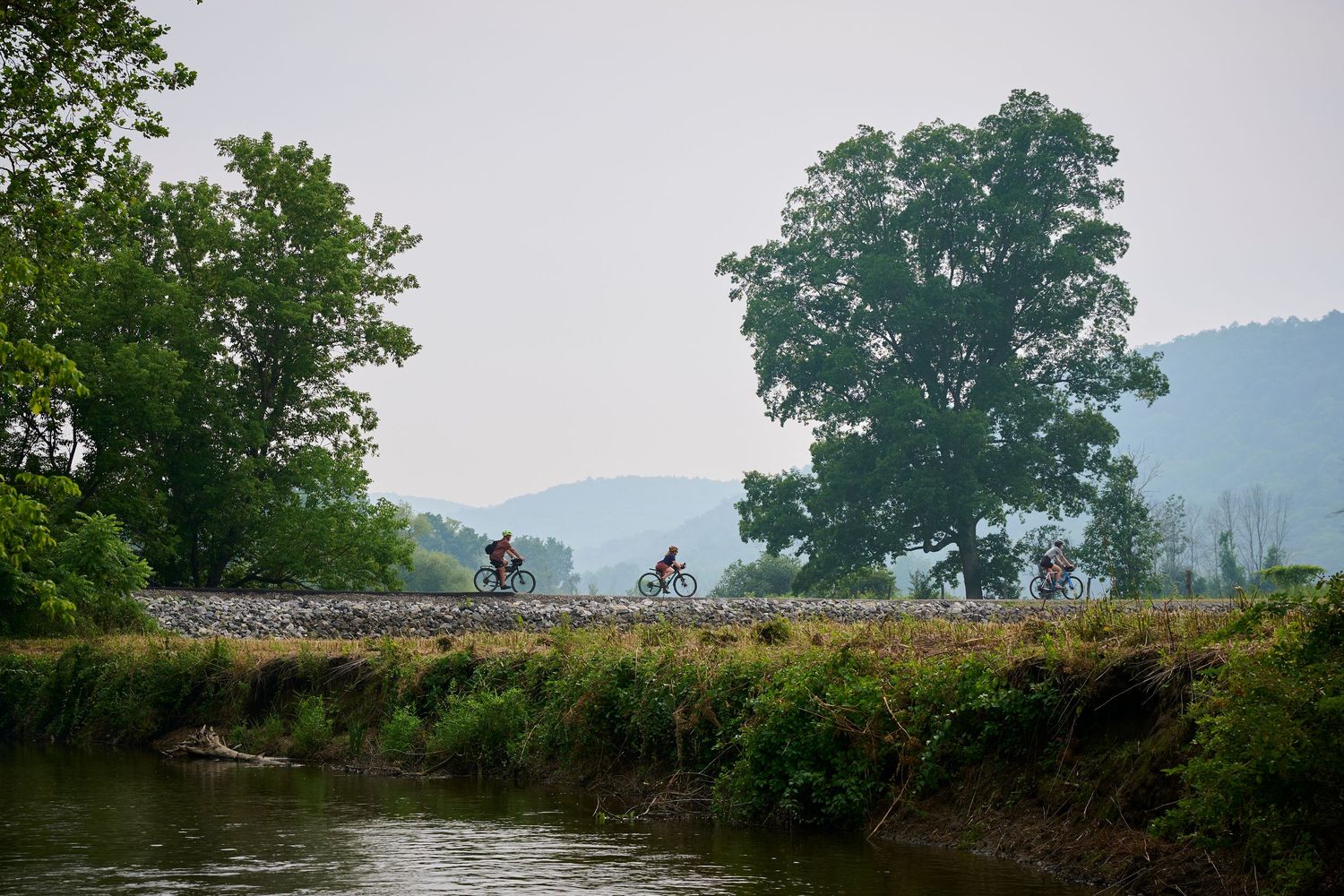 Cyclists riding along a scenic dirt path through lush green countryside with trees and mountains in the background.