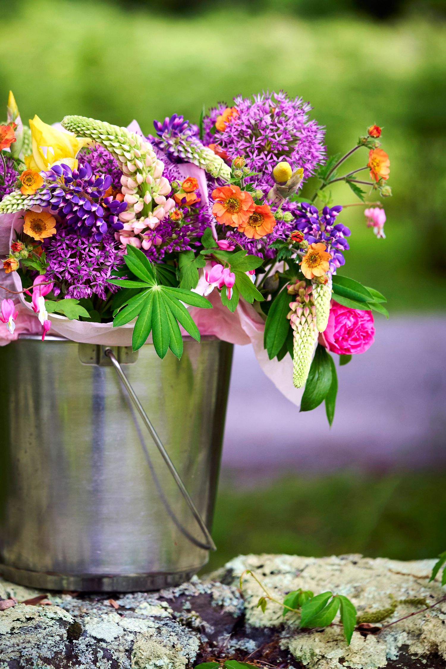 Colorful summer bouquet of purple allium, pink roses, and orange flowers arranged in a rustic metal bucket outdoors.