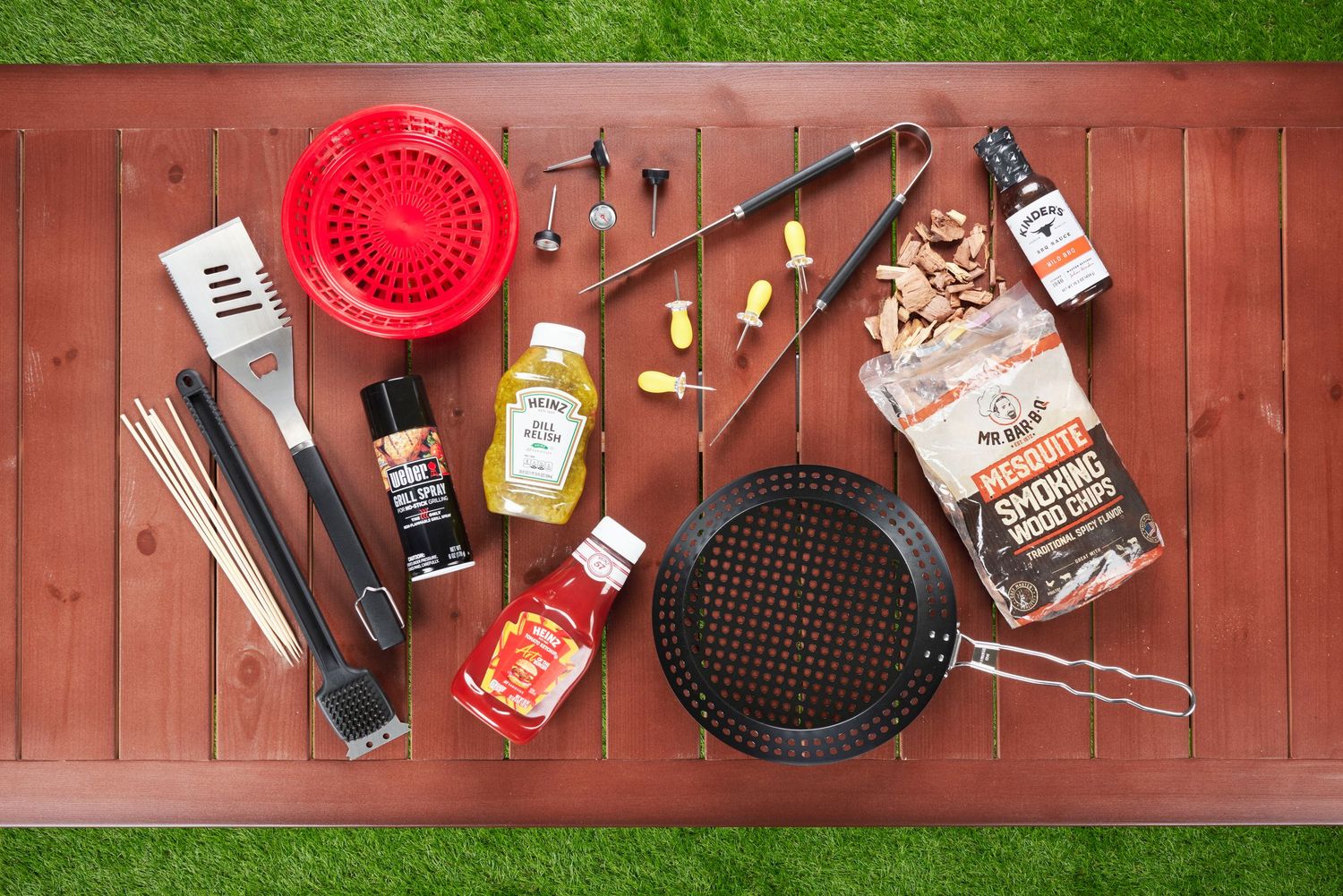 BBQ grilling accessories flatlay including spatula, brush, wood chips, sauces, and skewers on a wooden table.