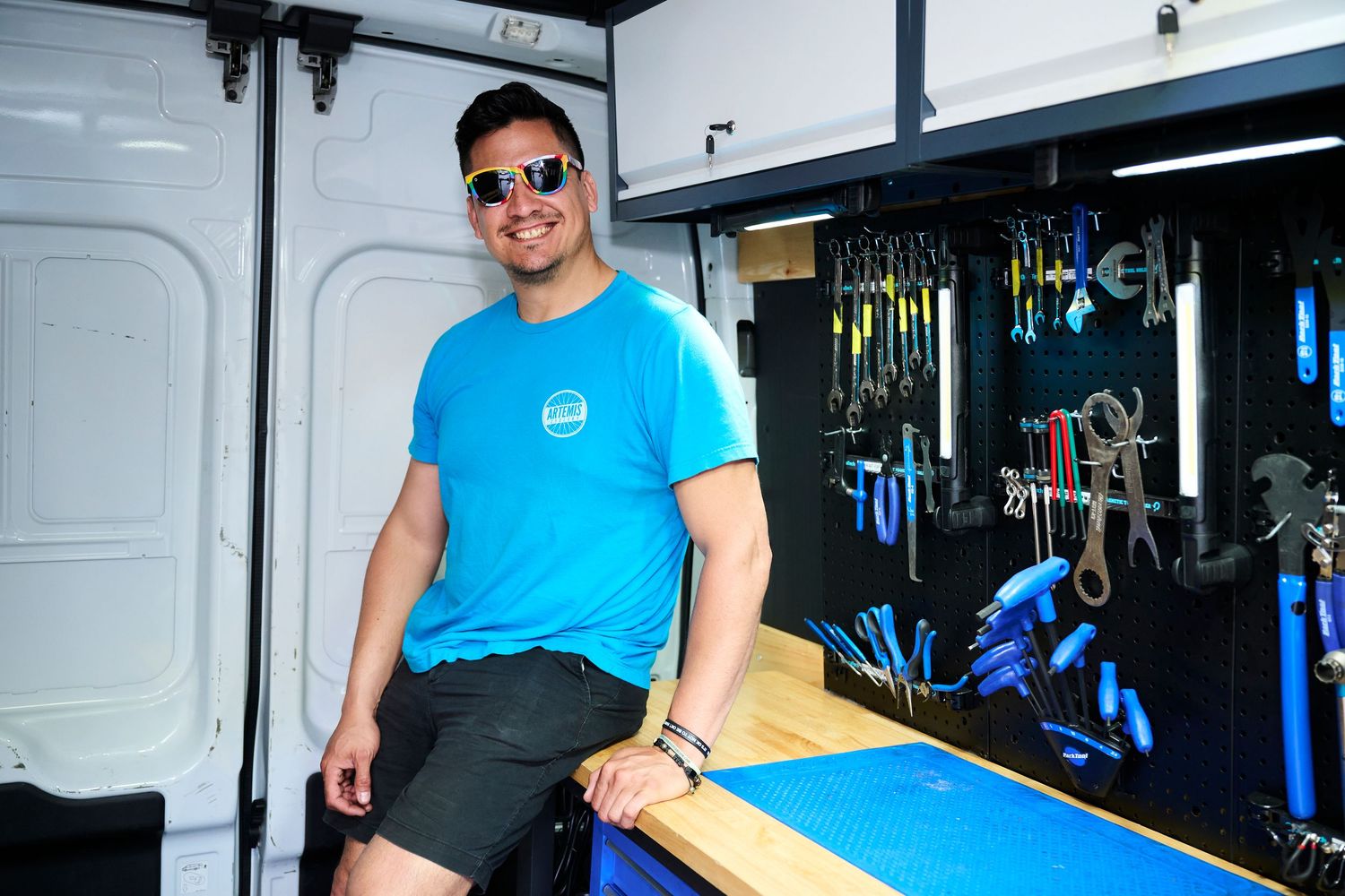 Man in blue t-shirt and sunglasses sitting in a mobile bike repair van with tools organized on the wall.