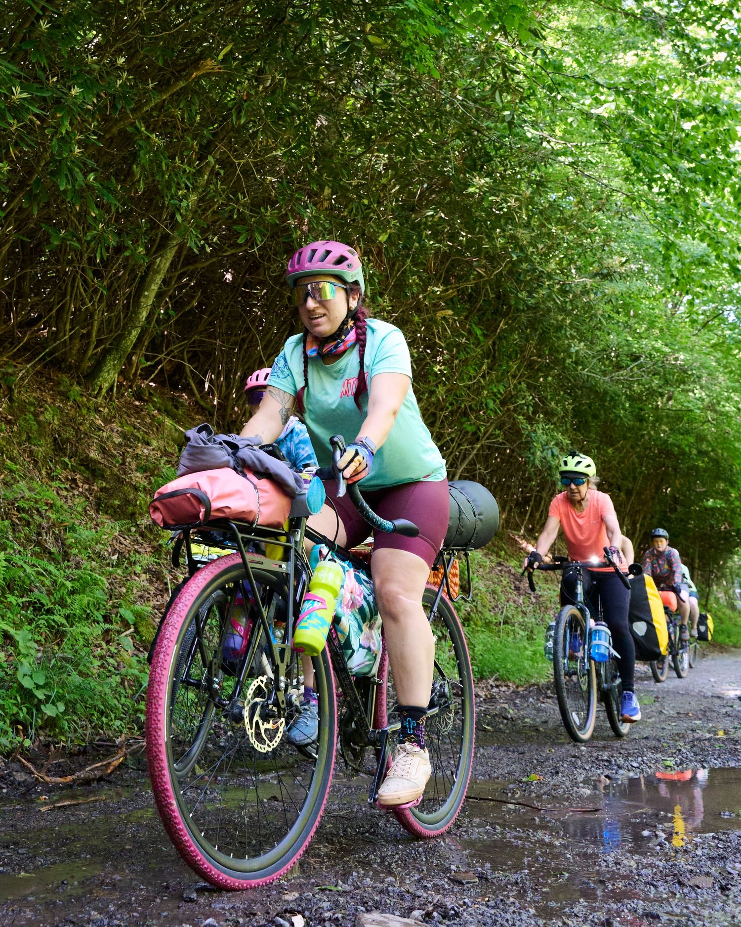 Group of cyclists bikepacking on a gravel trail through lush green forest, led by a woman in pink helmet with loaded panniers.