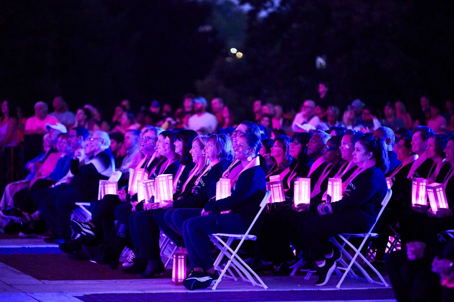 Audience members holding glowing candles at an outdoor evening ceremony illuminated by purple and pink lights.