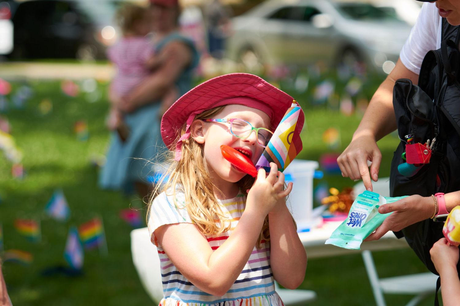 Young girl with face paint and red hat enjoys outdoor Pride festival event on sunny day.