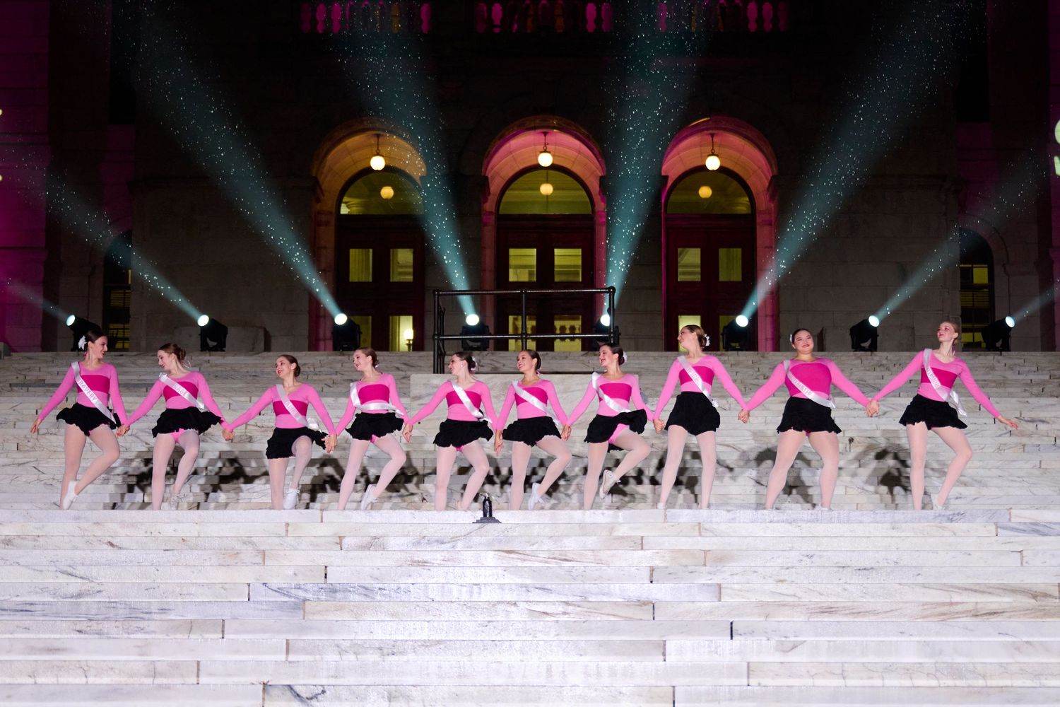 Row of silhouettes against pink lighting on outdoor stage steps.