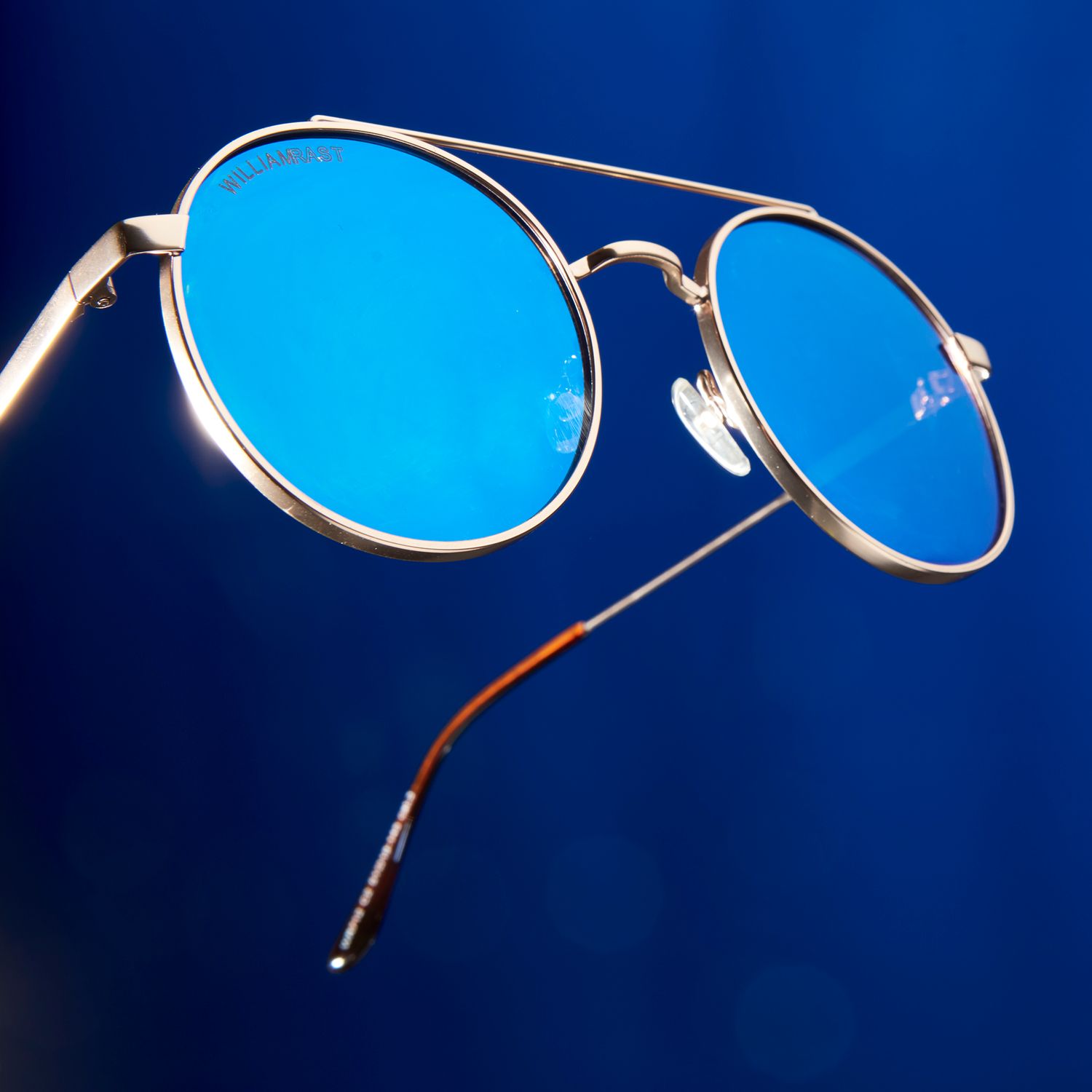 Round aviator sunglasses with blue mirrored lenses and gold metal frames against a deep blue background.