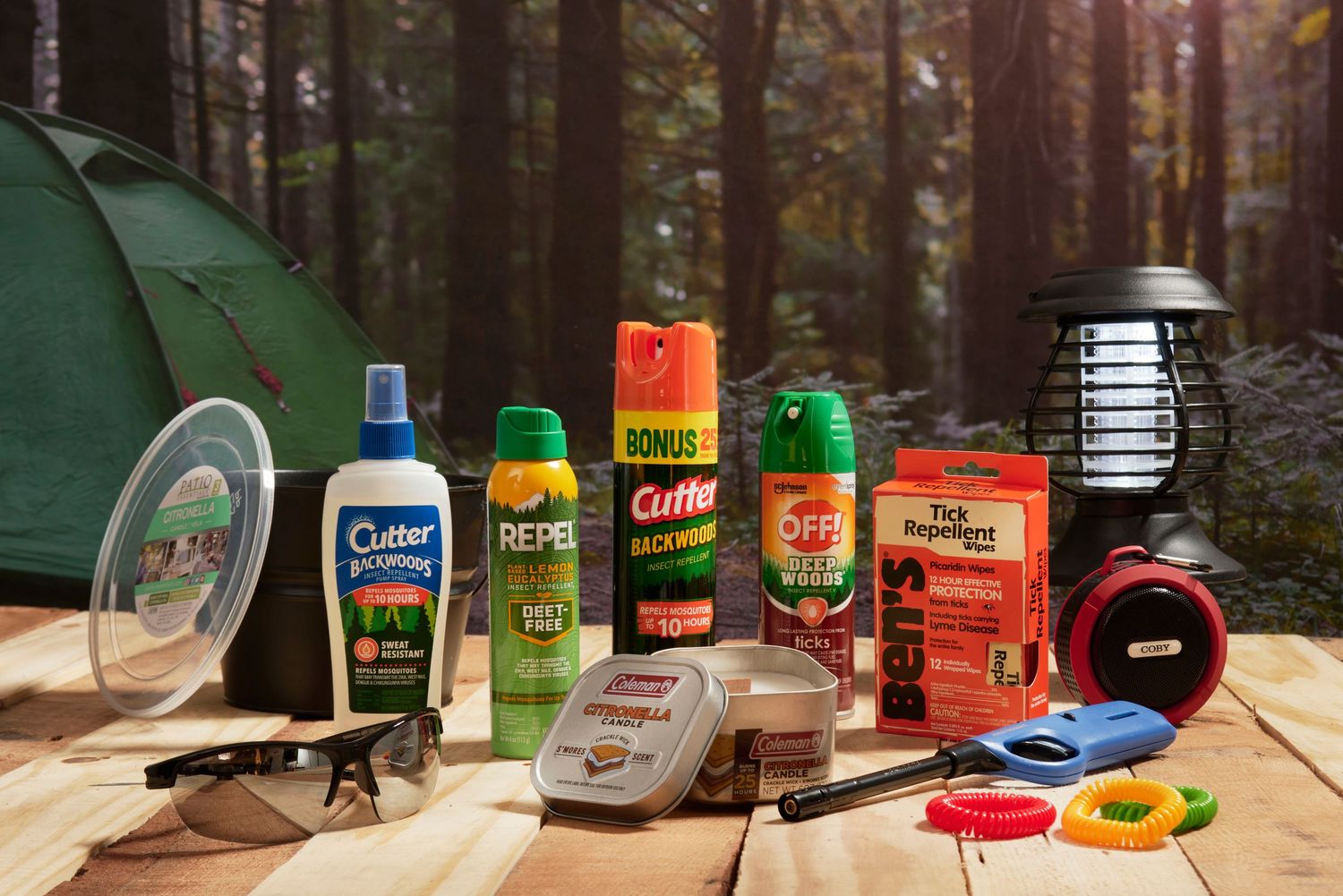 Camping essentials including Cutter, Repel, and OFF! insect repellents, a bug zapper lantern, and gear on a wooden table.
