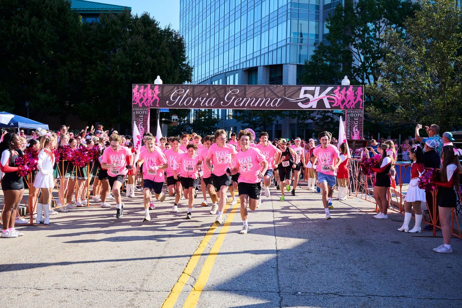 Runners in pink shirts start the Gloria Gemma 5K race on a sunny day with a pink finish arch.