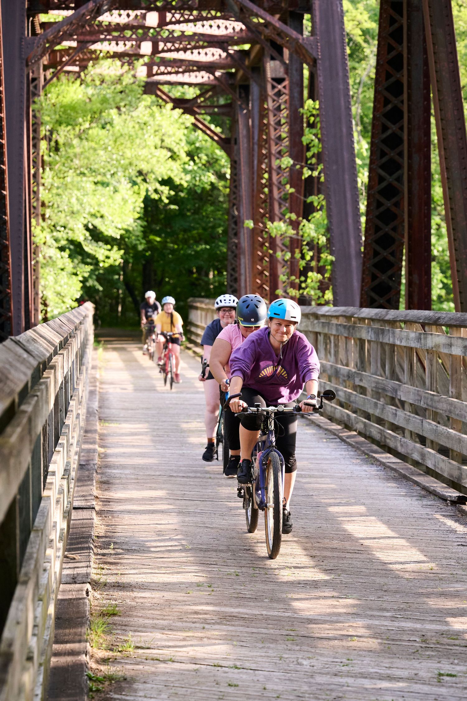 Group of cyclists riding bikes across a rustic iron truss bridge surrounded by lush green trees on a sunny day.