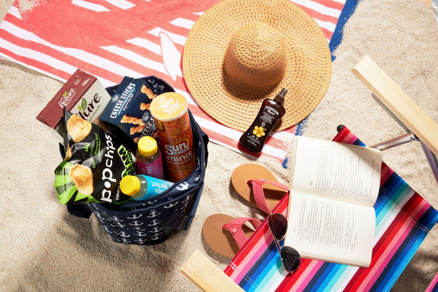 Beach essentials flat lay with snacks, sunscreen, hat, flip flops, sunglasses, and open book on colorful towel.