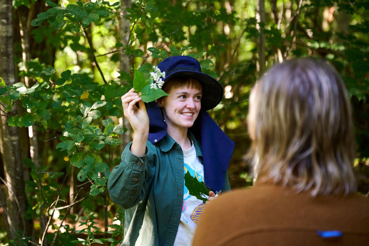 Woman in blue hat holds green leaf in sunlit forest, smiling at another person during outdoor nature walk.