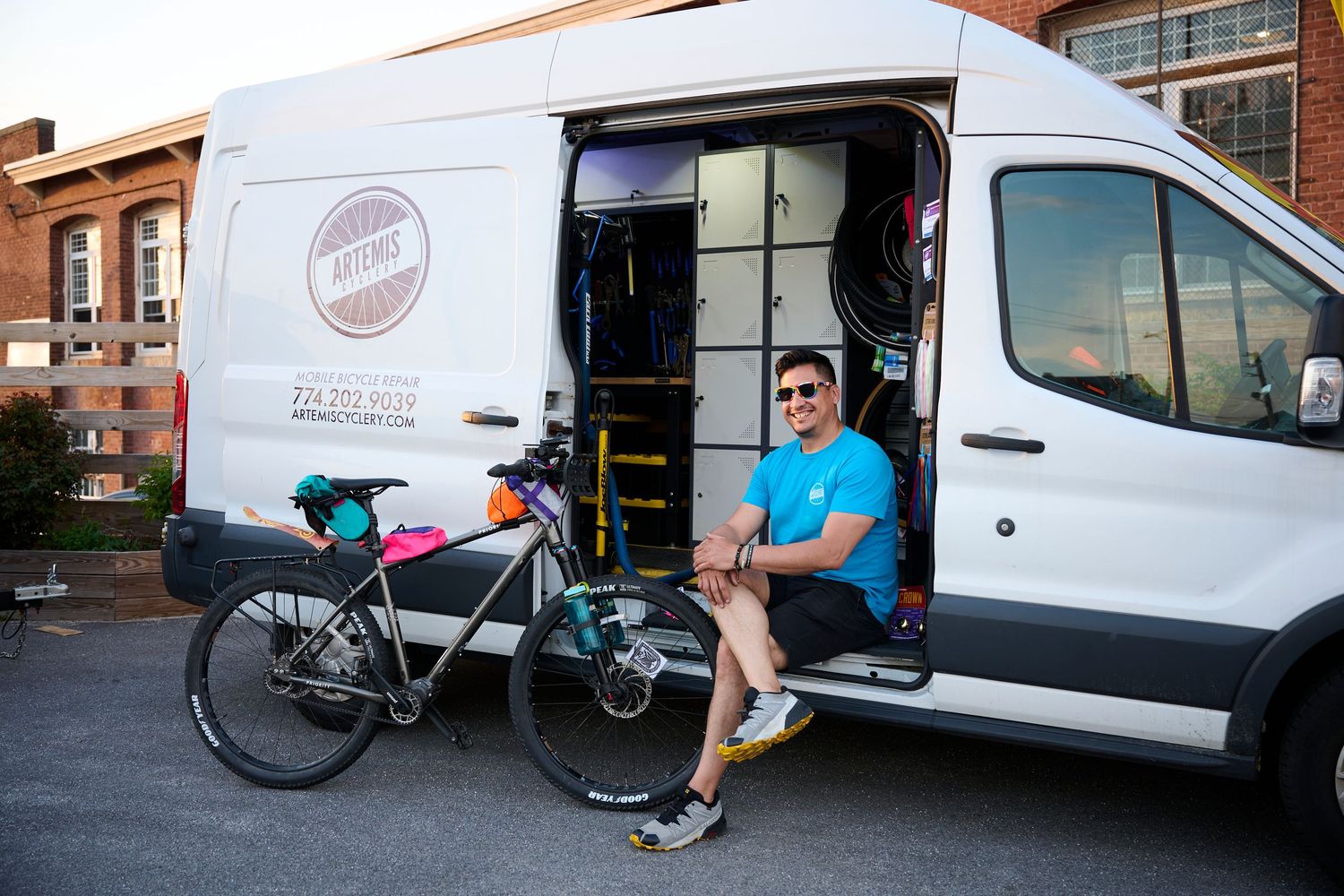 Man sitting in open white van next to a touring bicycle outdoors on a sunny day.