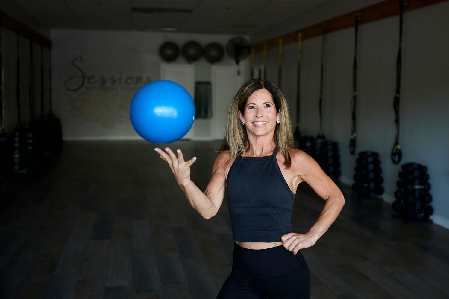 Woman in black athletic wear balancing a blue exercise ball on her finger in a fitness studio.