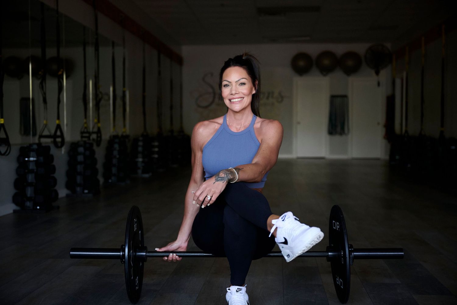 A smiling woman in athletic wear sits on a barbell in a gym, posing confidently among weights and fitness equipment.