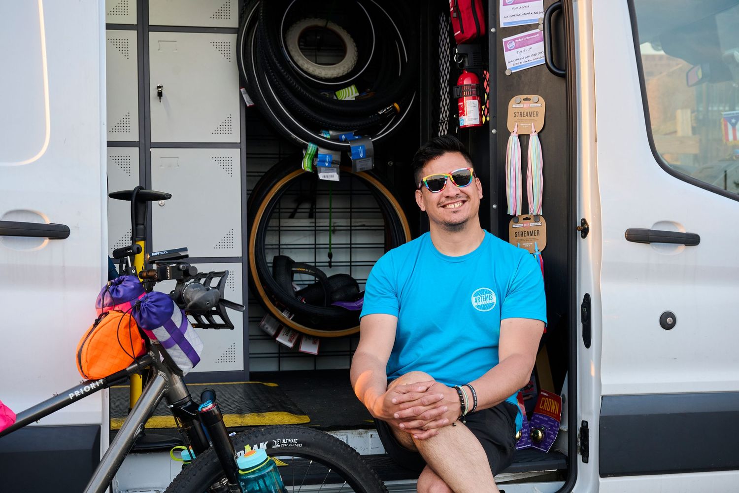 A mobile bike repair van with wheels mounted inside and someone sitting in the doorway wearing a bright blue shirt and sunglasses.