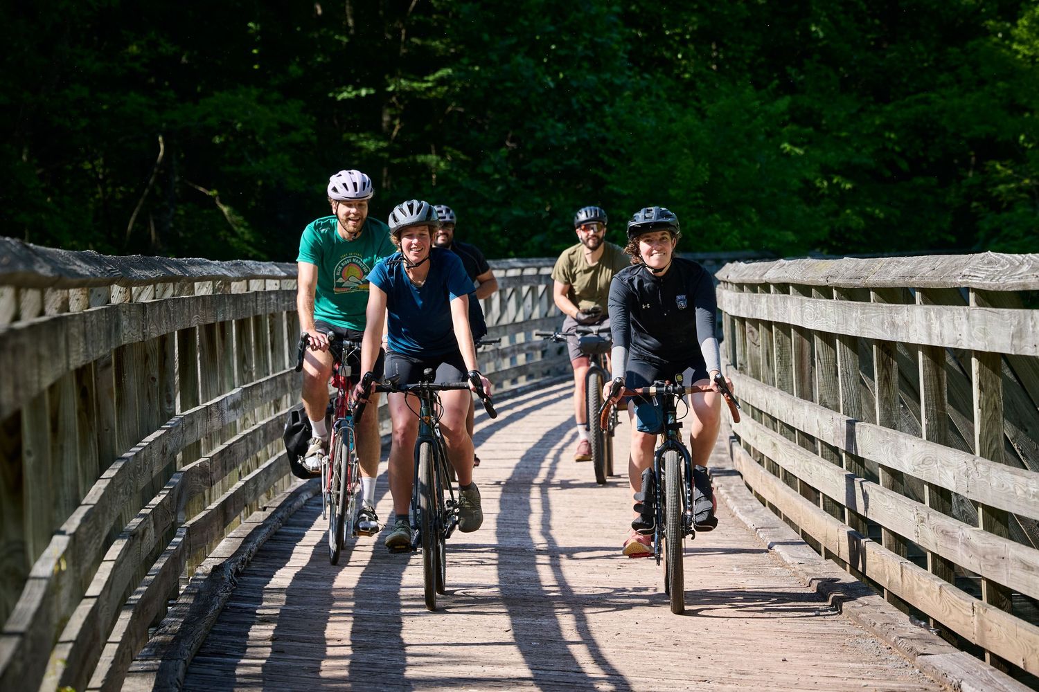 Four cyclists riding bikes across a wooden bridge surrounded by lush green trees on a sunny day.