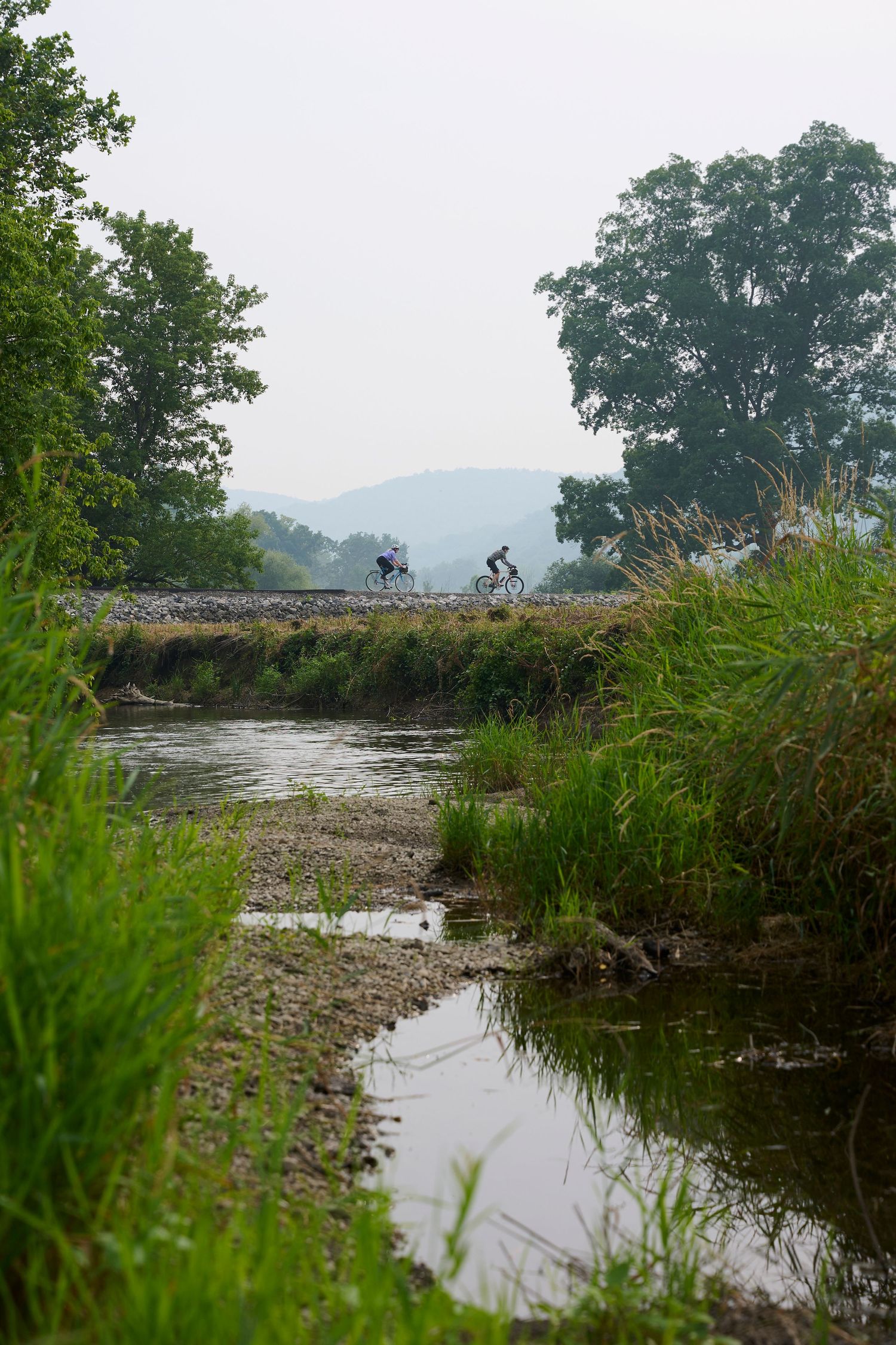 Distant cyclist silhouetted against misty mountains beside a winding stream.