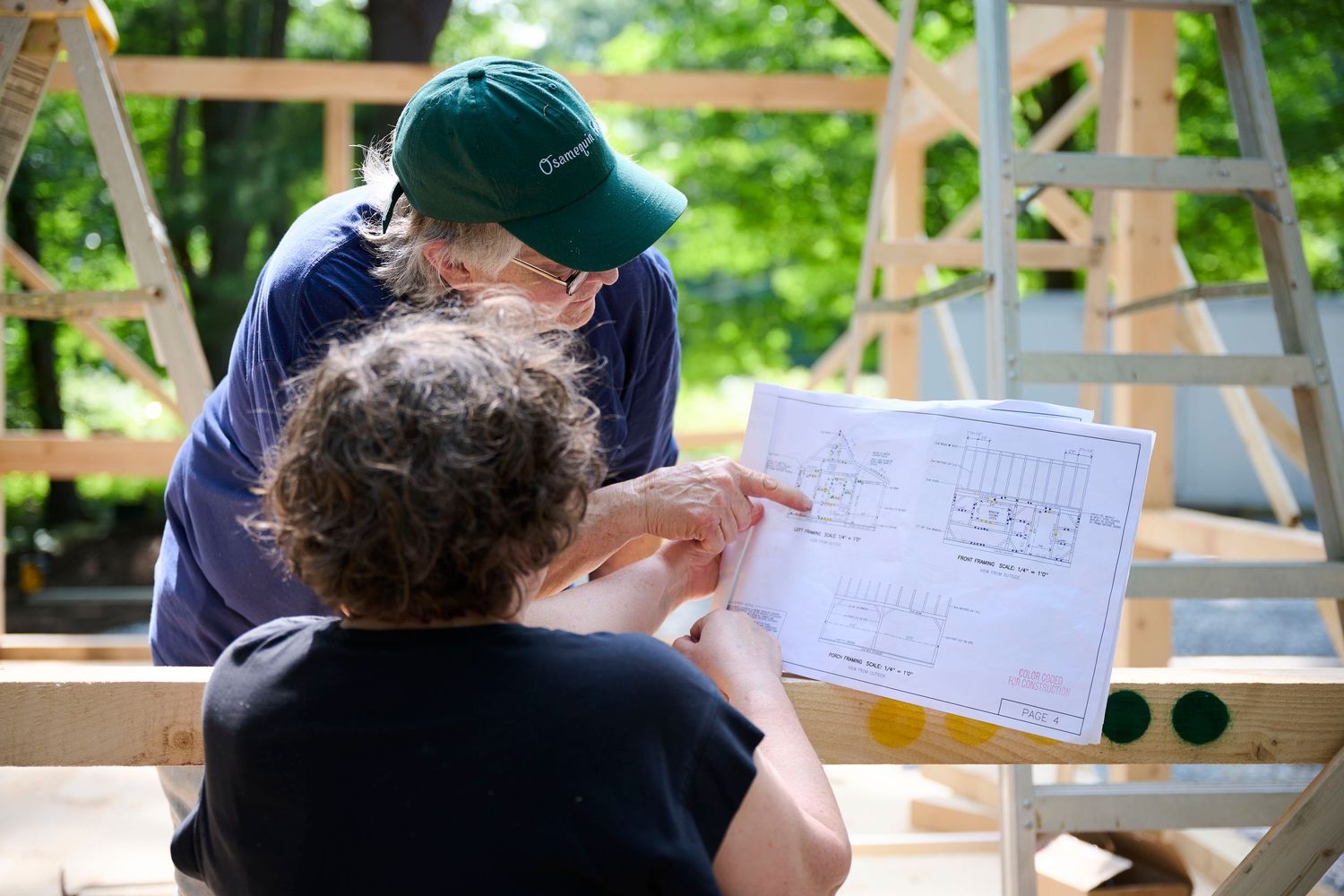Two construction workers reviewing building blueprints at a wooden framing site surrounded by trees.