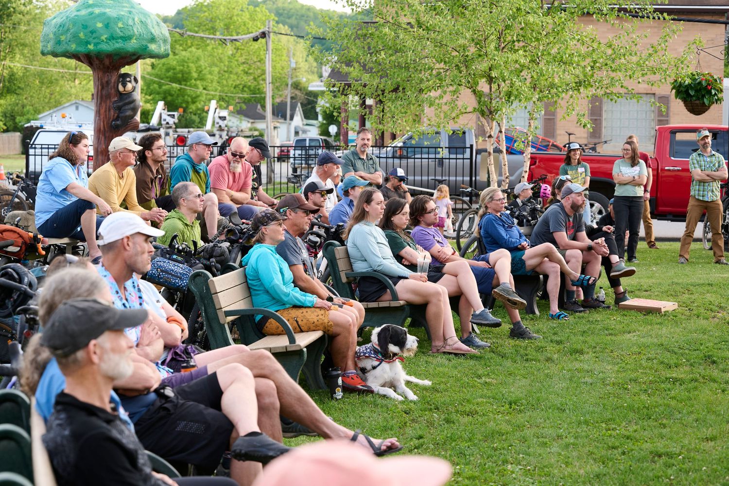 Crowd of people sitting on benches and grass at an outdoor community event on a sunny day with a dog nearby.