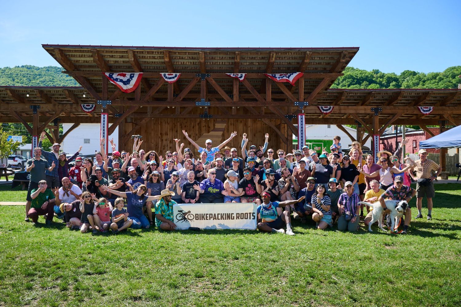 Large group of backpacking enthusiasts posing together in front of a rustic wooden pavilion at an outdoor festival.