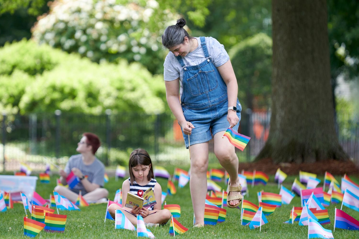 Adult and child placing rainbow Pride flags on green grass in a sunny park during an outdoor LGBTQ+ celebration event.