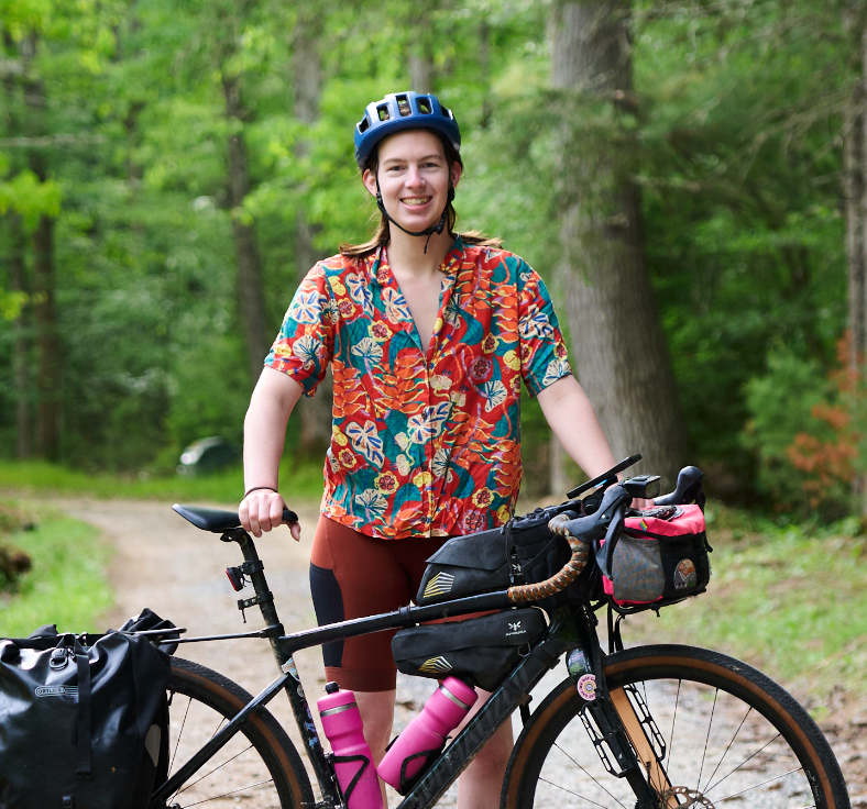 Smiling cyclist in colorful floral shirt and blue helmet standing with a loaded gravel bike on a forest trail.
