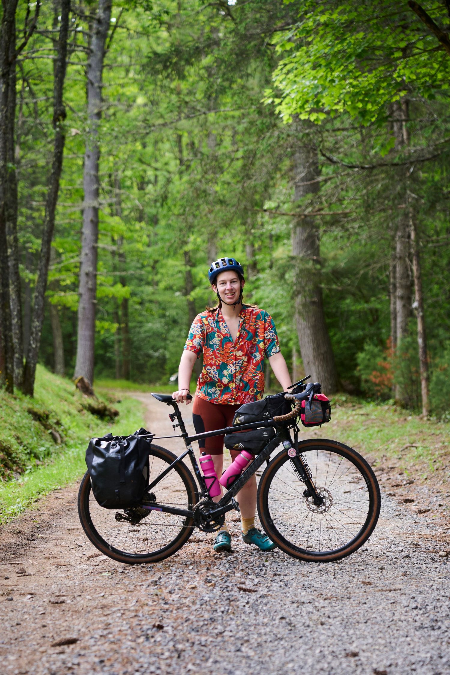 Smiling female cyclist in colorful shirt and helmet stands with loaded touring bike on gravel forest path.