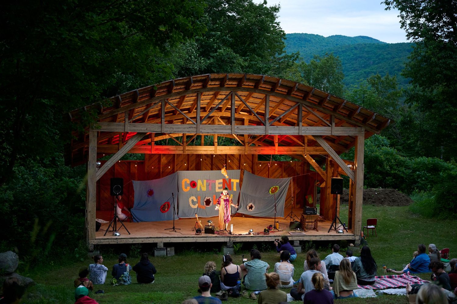 Outdoor theater performance at dusk with audience seated on grass, wooden stage, and mountain backdrop.