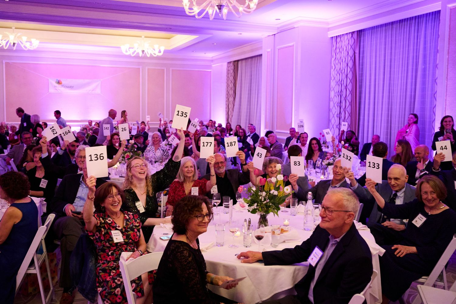 Guests raise numbered bidding paddles at a charity auction gala dinner in an elegant ballroom with purple lighting.