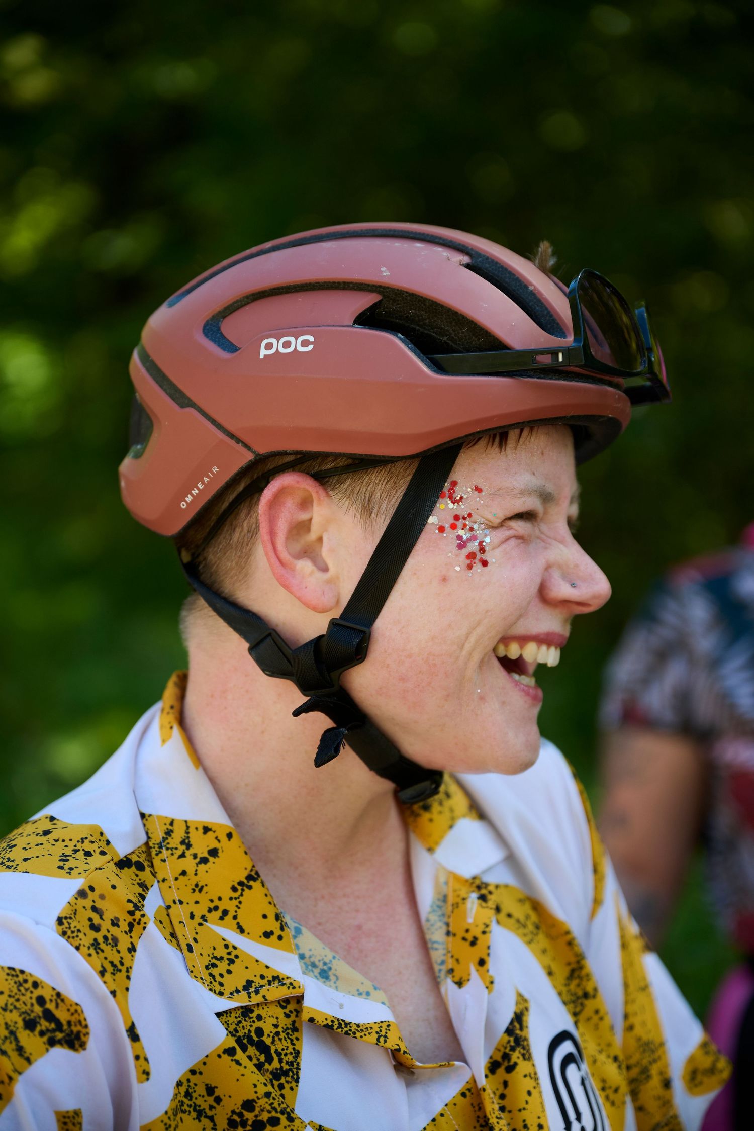 Smiling cyclist wearing a POC helmet with colorful face gems, dressed in a yellow patterned jersey outdoors.