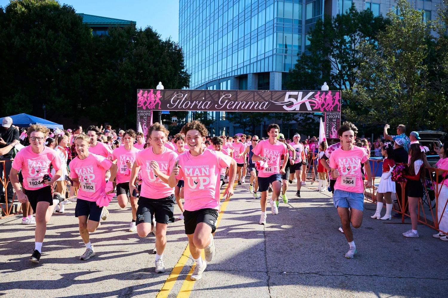 A group of participants wearing pink shirts gather at a charity walk event in an urban setting with tall buildings.