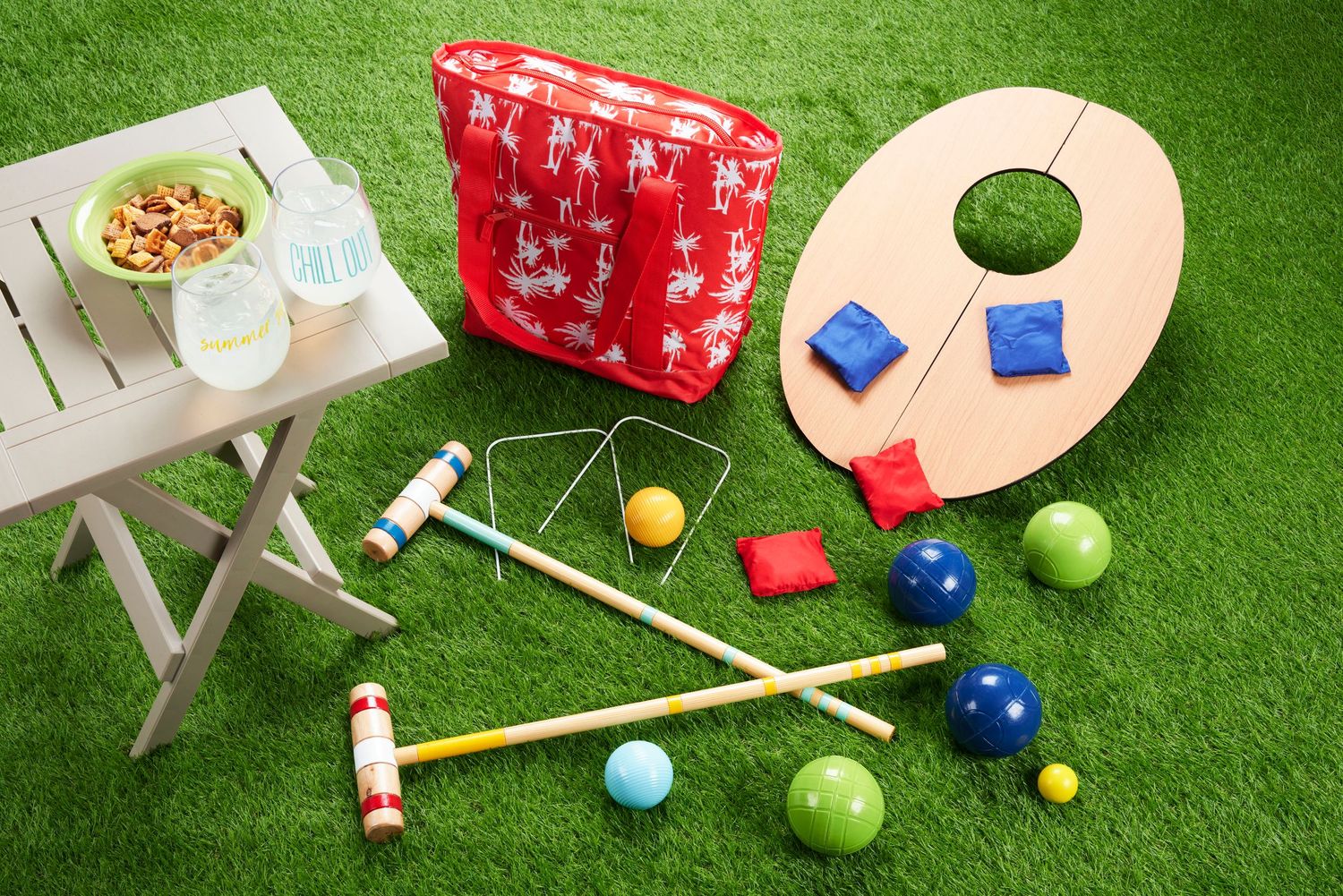 Outdoor lawn games set with croquet mallets, colorful balls, cornhole board, bean bags, and a red tote bag on green grass.