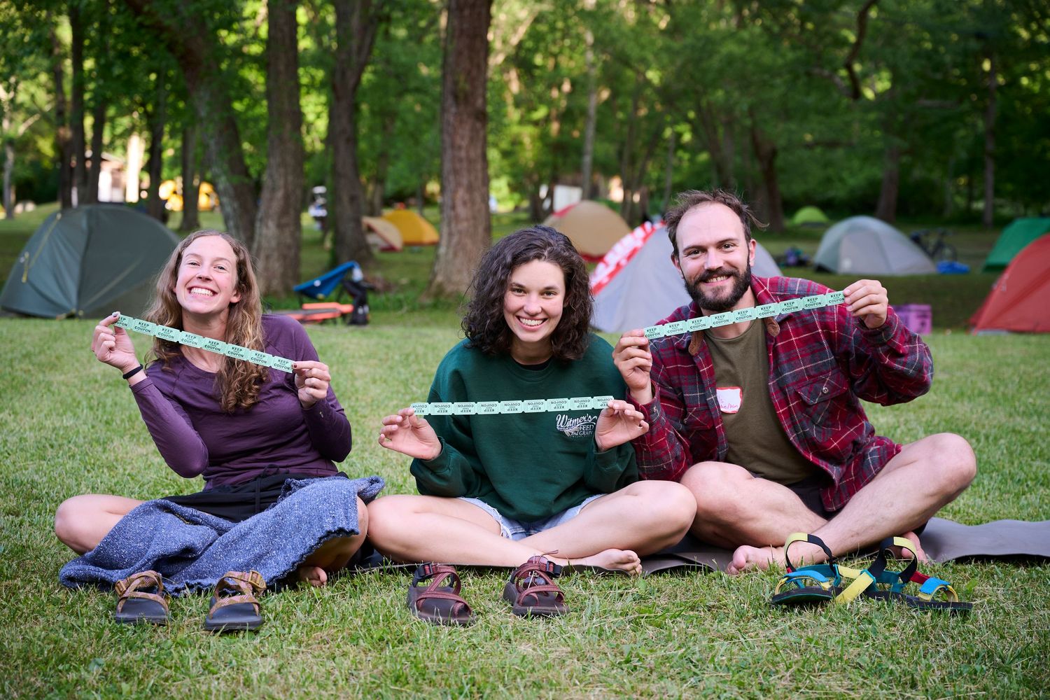Three friends sit on grass at a campsite, smiling and holding a green measuring tape together.